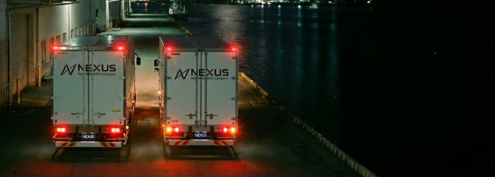 Two NEXUS trucks side by side at the port at night, lights on.